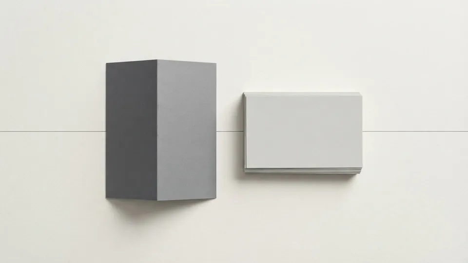 A folded grey paper card standing upright beside a small stack of blank index cards, a fine graphite rule running behind them